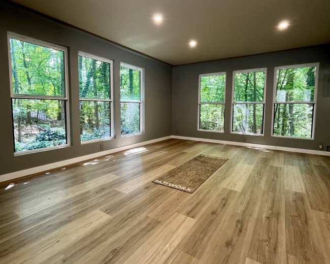 The image shows a spacious, empty room with large windows overlooking a wooded area, and light-colored wood flooring.