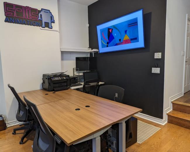 The image shows a small office space with a wooden table, four black chairs, a printer, and a colorful abstract artwork displayed on a wall-mounted screen.