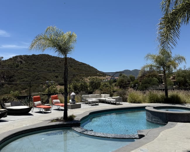 A swimming pool with a curved edge is surrounded by lounge chairs and palm trees, set against a backdrop of rolling hills and a clear blue sky.
