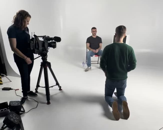 A film crew sets up in a studio with a seated interviewee in a chair, while a person kneels on the floor facing the camera.