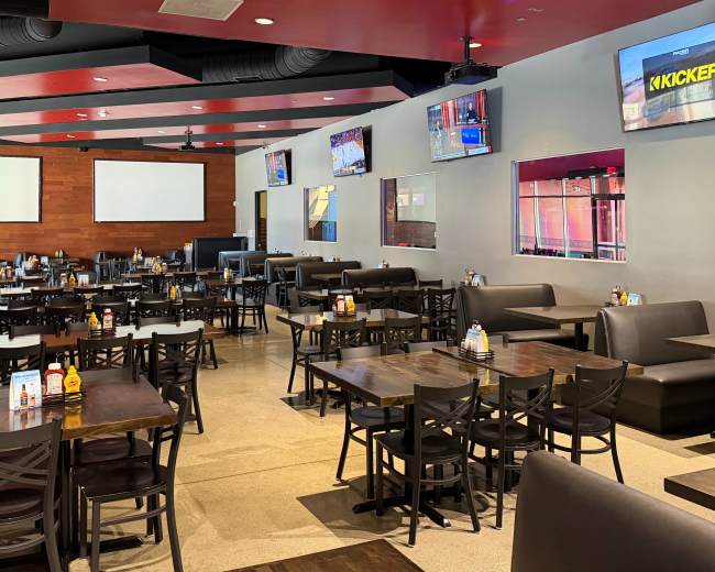 The image shows an interior of a restaurant with numerous tables and chairs arranged in a spacious layout, featuring multiple televisions mounted on the walls.