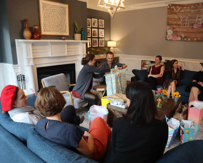 A group of people sits in a living room with presents and decorations, while one person unwraps a gift.