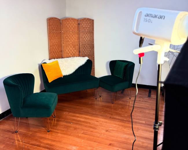 The image shows a cozy photography studio with a green velvet sofa and chairs, a wicker screen in the background, and a lighting setup on the right.