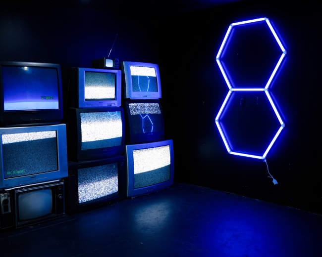 A collection of vintage televisions displaying static is arranged against a dark wall, alongside a neon blue hexagonal light fixture forming the number 8.