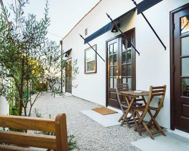 The image shows a narrow pathway lined with olive trees, leading to several doors with dark wooden frames and a small outdoor table with chairs.