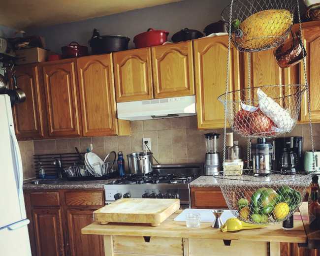 A kitchen features wooden cabinets, a countertop island with a basket of fruit, and various appliances on the counters.