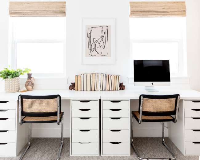 A modern home office features two desks with matching chairs, surrounded by storage drawers and illuminated by natural light from two windows with shades.