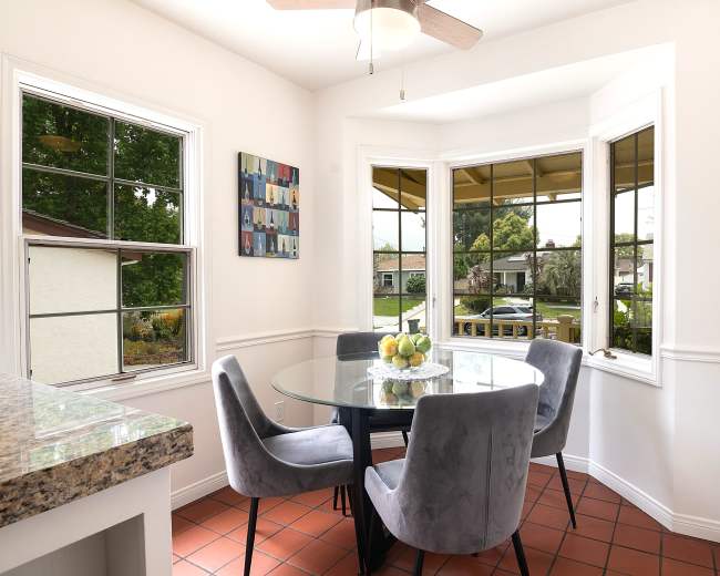 A round dining table with gray chairs is positioned near large windows in a bright kitchen area, showcasing a view of the outdoors.