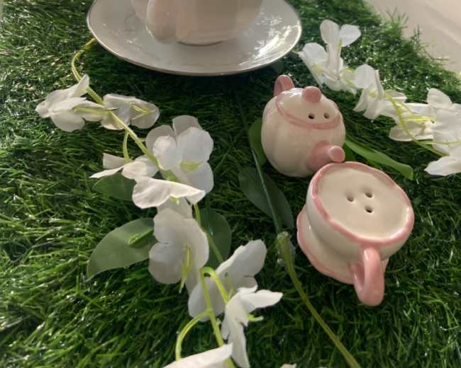 The image shows a table setting with a teapot, an empty cup on a saucer, and decorative floral elements on a green grass-like table runner.