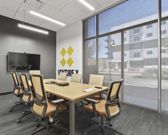A modern conference room features a large table surrounded by eight ergonomic chairs, a black screen mounted on the wall, and a logo displayed on the gray backdrop.