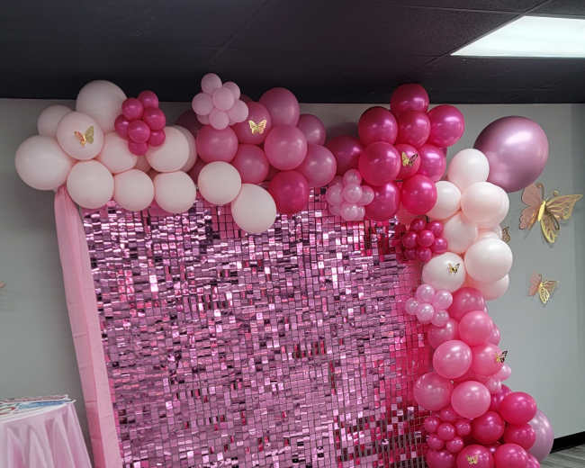 The scene features a backdrop made of pink balloons and shiny decorations, alongside a pink tablecloth draped over a table.