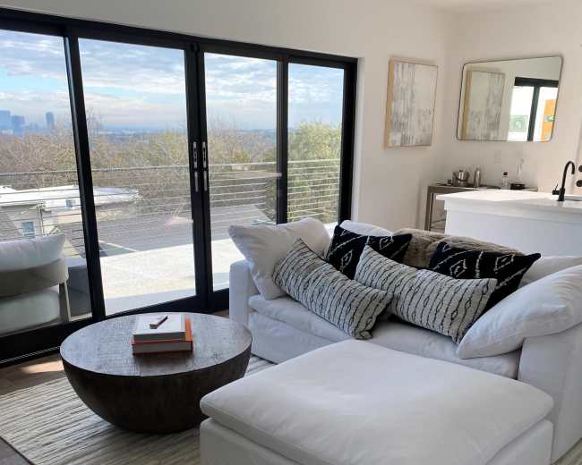 A bright living room features a white sectional sofa with black and white patterned pillows, a round wooden coffee table, and a large window offering a view of the city skyline.