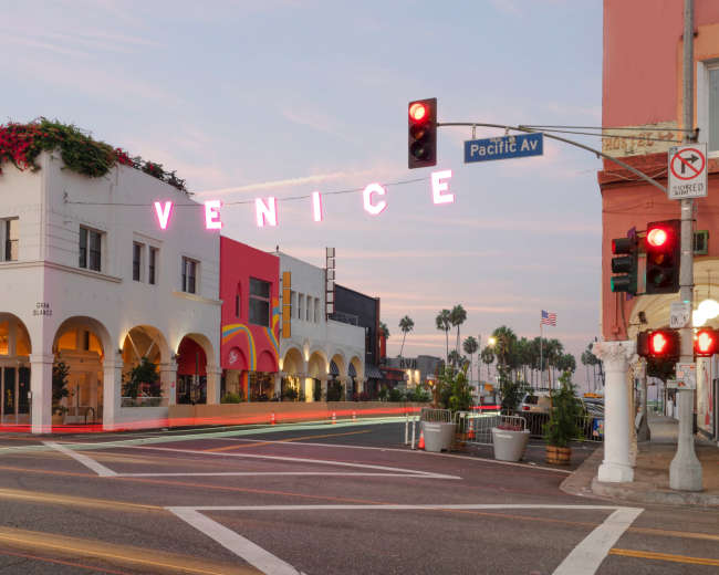The image shows a street scene in Venice, California, featuring buildings with vibrant facades and a neon sign that reads "VENICE" above the intersection at Pacific Avenue.