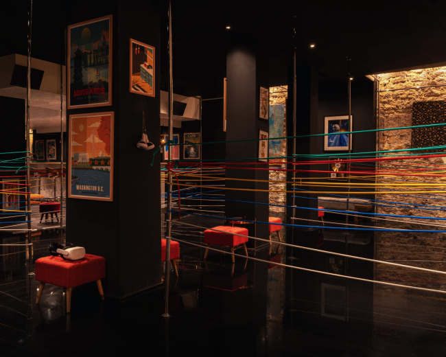 The image depicts an art installation featuring colorful lines stretched across a dark room with red stools and framed posters on the walls.