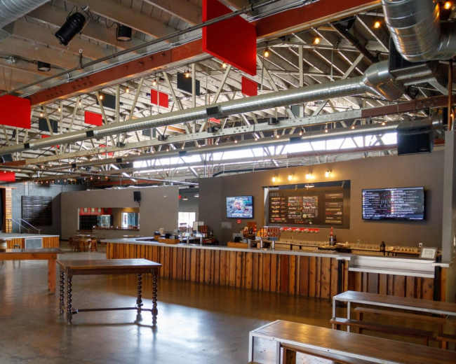 A spacious, modern interior of a venue with high ceilings, wooden tables, and a bar area featuring screens and a menu board.