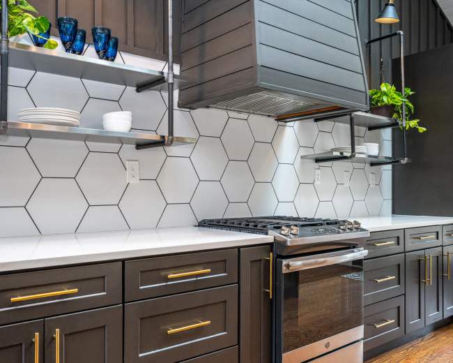 The image shows a modern kitchen with black cabinets, a stainless steel oven, and hexagonal tile backsplash, complemented by open shelving displaying plants and tableware.