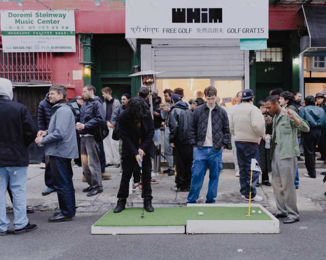 A crowd gathers outside a storefront while two people play mini-golf on a small green setup in the street.