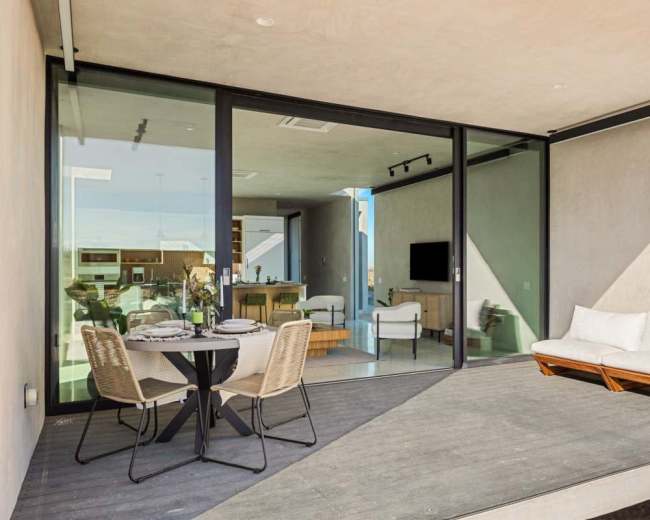 A modern patio features a small dining table with chairs and a sofa, enclosed by large glass sliding doors.