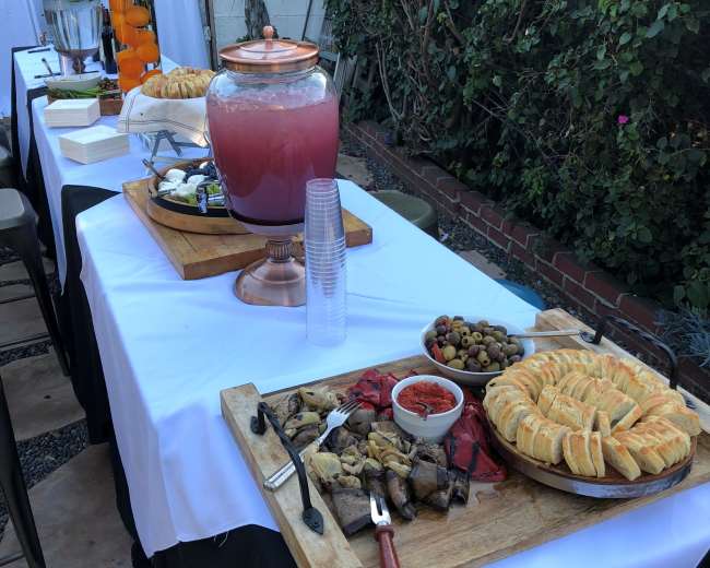 A table is set with various food items, including pastries, olives, and a drink dispenser, surrounded by greenery.