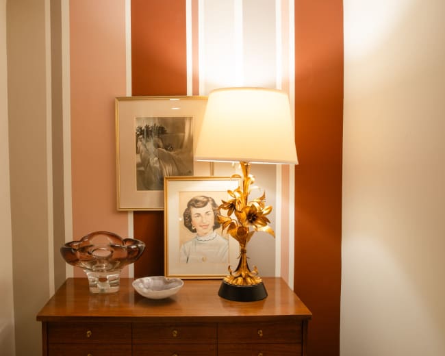 A wooden dresser displays a lamp with a floral design, a glass vase, and two framed pictures against a striped wall.