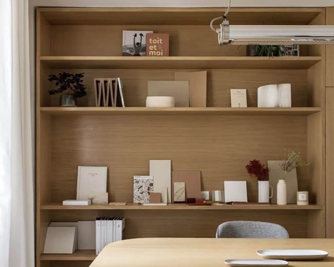 The image shows a minimalist wooden desk in front of a neatly organized shelf filled with various decorative items and stationery.