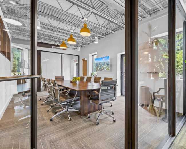 A modern conference room features a large table surrounded by chairs, with glass walls, a television, and plants in the background.