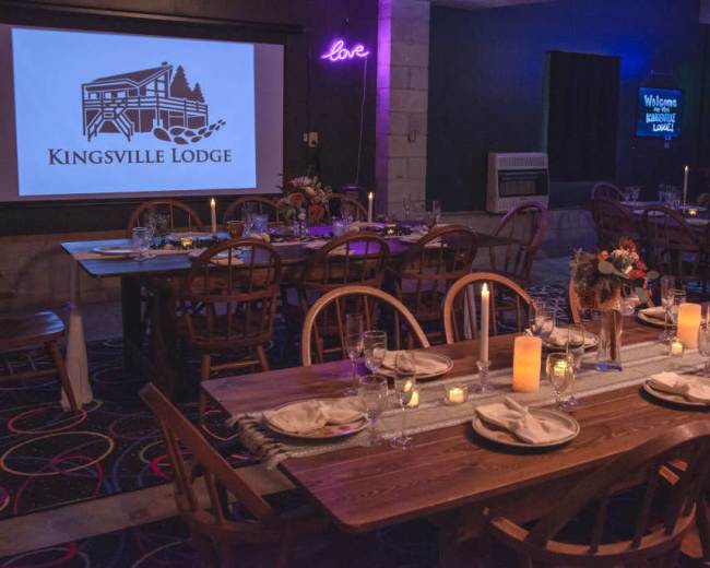 The image shows a dining area with wooden tables set for a meal, featuring candles and flower centerpieces, alongside a logo of "Kingsville Lodge" displayed on a screen.