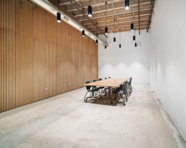 The image shows a spacious meeting room with a long wooden table surrounded by rolling chairs, and wooden paneling on one wall, illuminated by overhead lights.