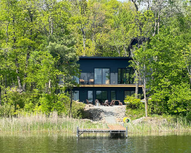 The image shows a modern black house overlooking a calm lake, surrounded by lush greenery and a wooden dock extending into the water.
