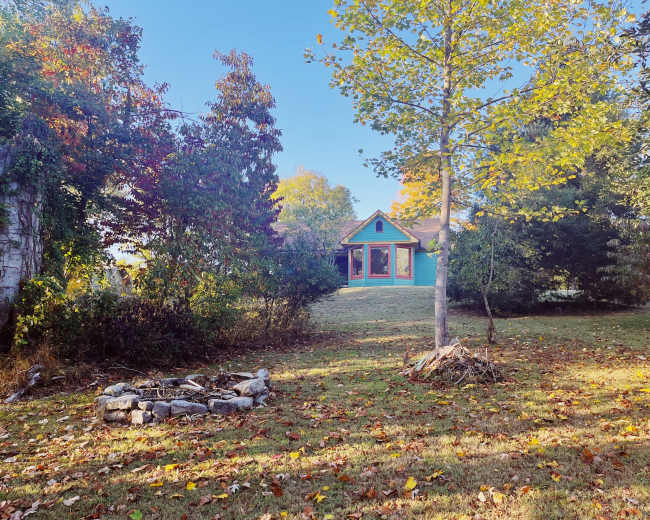 A small blue house is set in a yard surrounded by trees and colorful autumn foliage, with a stone fire pit in the foreground.