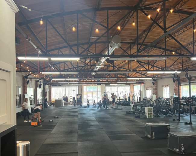 The image shows a spacious gym interior with black rubber flooring, weightlifting equipment, and several people engaged in workouts.