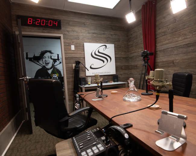 A recording studio with a wooden table, microphones, and a large clock displaying the time.