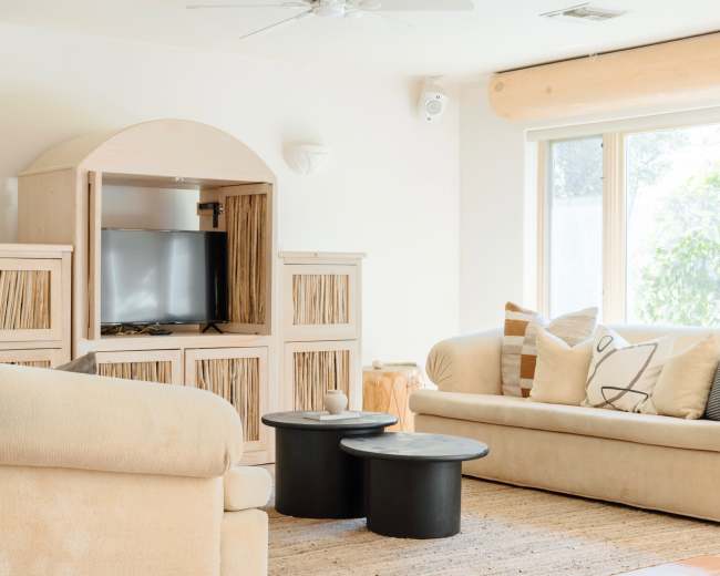 The room features two beige sofas, a wooden entertainment unit with a television, and two black circular coffee tables on a light woven rug.