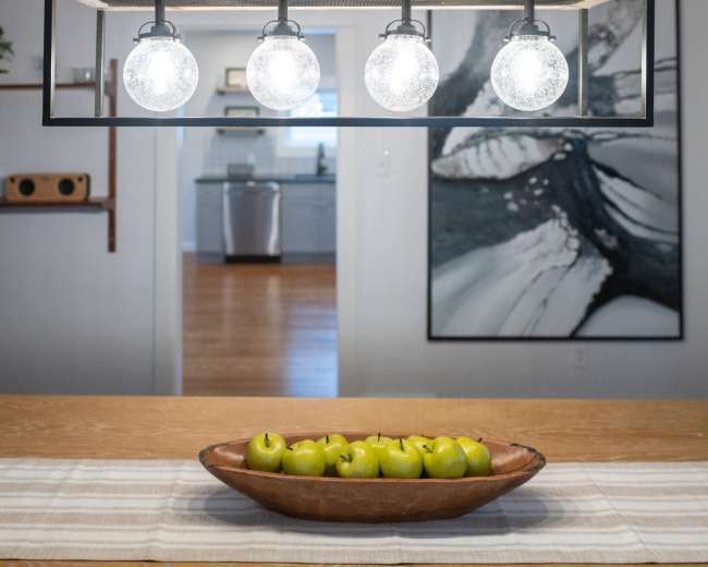A wooden dining table with a bowl of green apples is illuminated by a four-bulb pendant light hanging overhead.