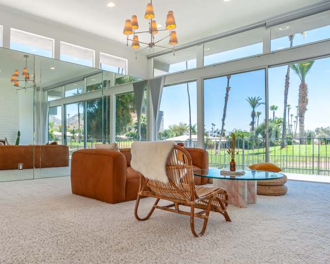 The scene shows a bright, modern living room with large glass windows, reflecting walls, and stylish furniture, including a leather couch and a rattan chair.