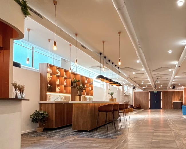 The image shows a modern interior space with wooden accents, featuring a long seating area, a bar-like counter, and overhead pendant lighting.