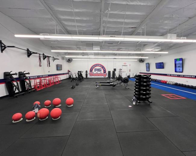 The image shows a gym interior with various fitness equipment, including weights, benches, and exercise balls arranged across a black rubber floor.