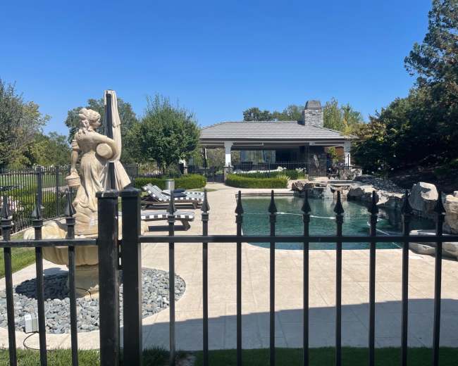 A swimming pool with a decorative statue stands in a landscaped backyard, adjacent to a covered porch and trees.