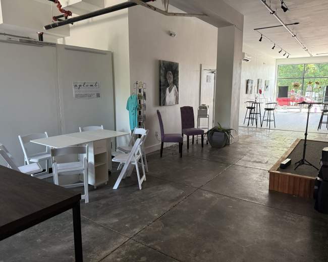 The image shows a spacious, minimalist indoor area with a white table and chairs, a purple chair, and artwork on the walls, leading to a large glass entrance.