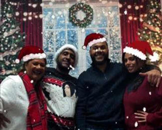 A group of four people poses in front of a window decorated for Christmas, with snowflakes falling and wearing Santa hats.