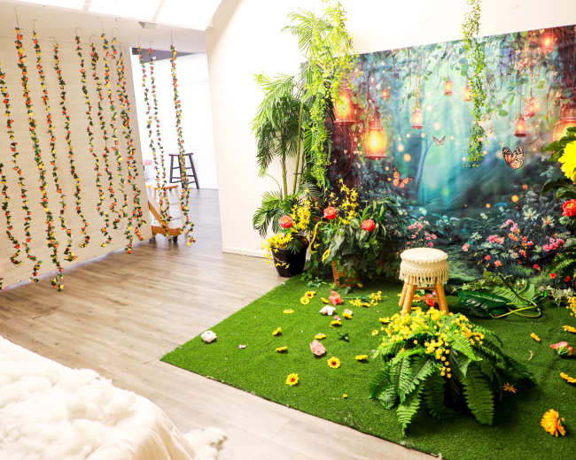 The image features a vibrant photo studio with lush green artificial grass, a floral backdrop with lanterns, and decorative plants arranged alongside a wooden stool and scattered flowers.