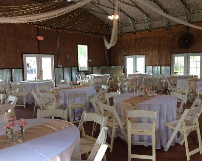 A spacious event venue with round tables covered in white tablecloths and pink decorative elements, surrounded by a wooden interior and string lights overhead.