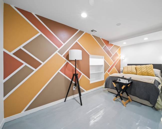 A modern bedroom with an abstract geometric accent wall, a bed with yellow and gray bedding, and a black floor lamp beside a small folding table.