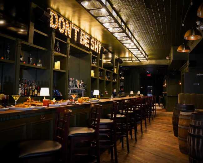 The image shows a dimly lit bar with a long counter, high chairs, shelves filled with various bottles, and wooden barrels along the walls.