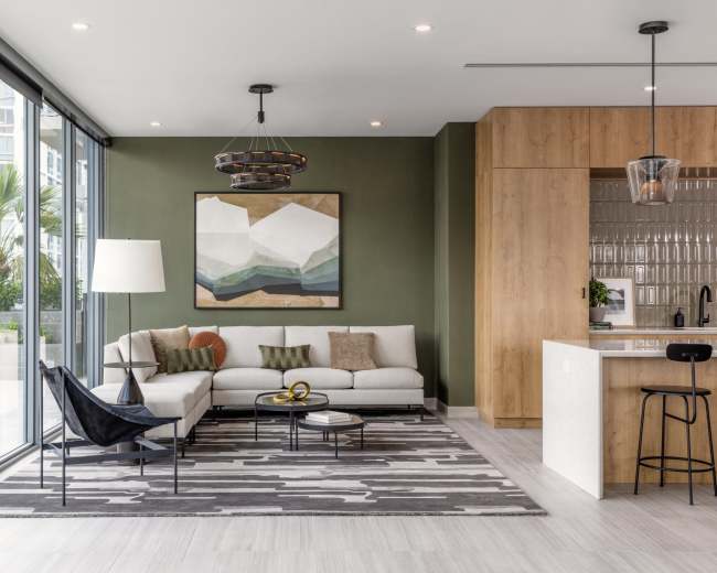 The image shows a modern living room with large windows, featuring a light-colored sofa, a geometric wall art piece, a round coffee table, and a kitchen area in the background.