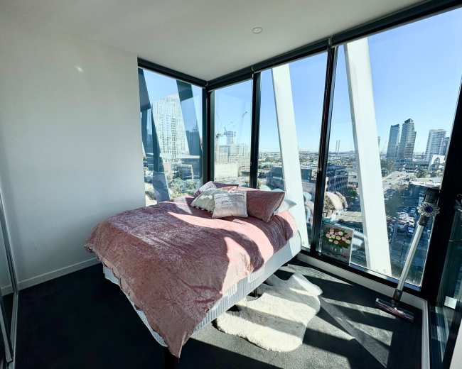 A modern bedroom with large glass windows showcasing a city skyline, featuring a bed with a pink blanket and a plush rug on the floor.