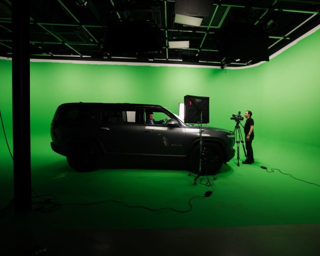 A gray SUV is positioned in a studio with a green backdrop, while a person operates a camera nearby.