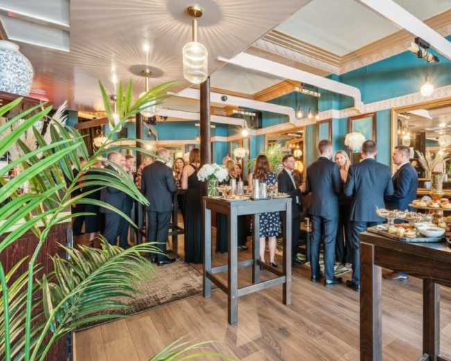 A group of well-dressed people mingles in a stylishly decorated event space, surrounded by food displays and plants.