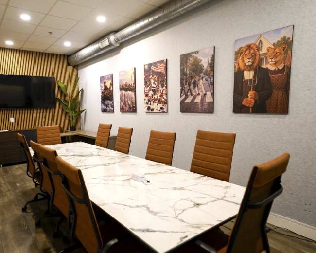 A modern conference room features a long marble table surrounded by leather chairs, with framed artwork on the walls and a television mounted in one corner.