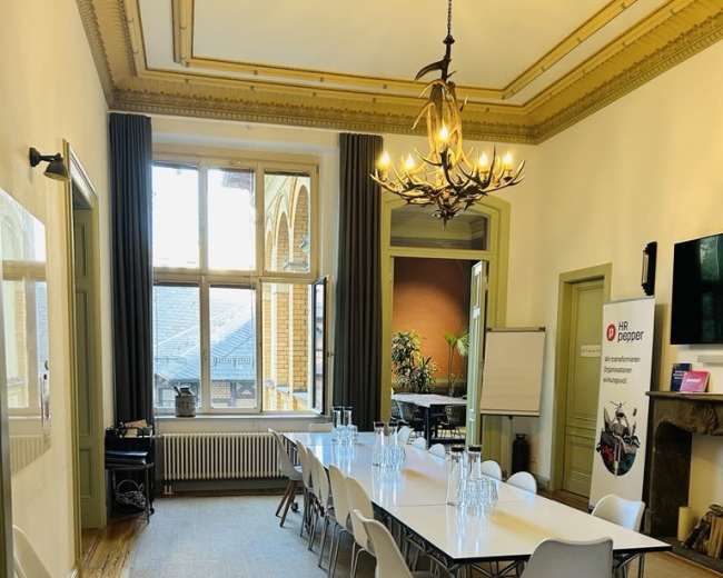 The image shows a spacious meeting room with a long table set for a presentation, featuring modern chairs, large windows, and decorative light fixtures.